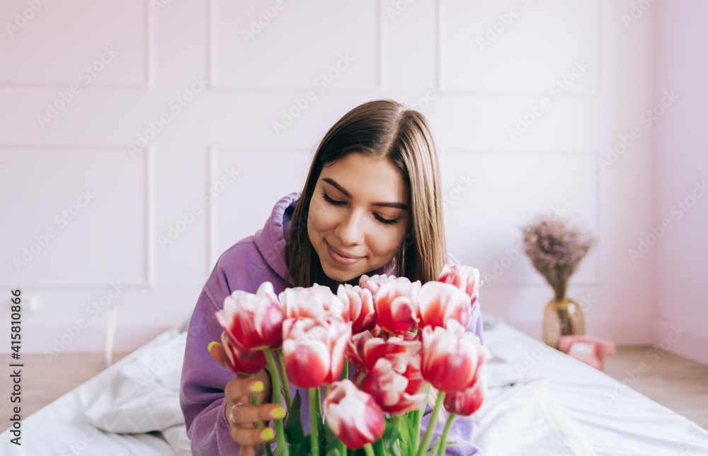 Young caucasian happy woman smelling bouquet of fresh tulips at home. Sitting on the bed in spring morning, woman day concept. Young caucasian happy woman smelling bouquet of fresh tulips at home. Sitting on the bed in spring morning, woman day concept.