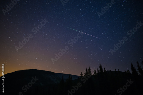 One meteor (shooting star) from the Perseids meteor shower on a summer night with a clear sky full of stars in the mountains with soft orange light from the city on the horizon, Kananaskis, Canada