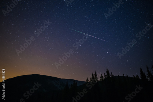 One meteor (shooting star) from the Perseids meteor shower on a summer night with a clear sky full of stars in the mountains with soft orange light from the city on the horizon, Kananaskis, Canada photo