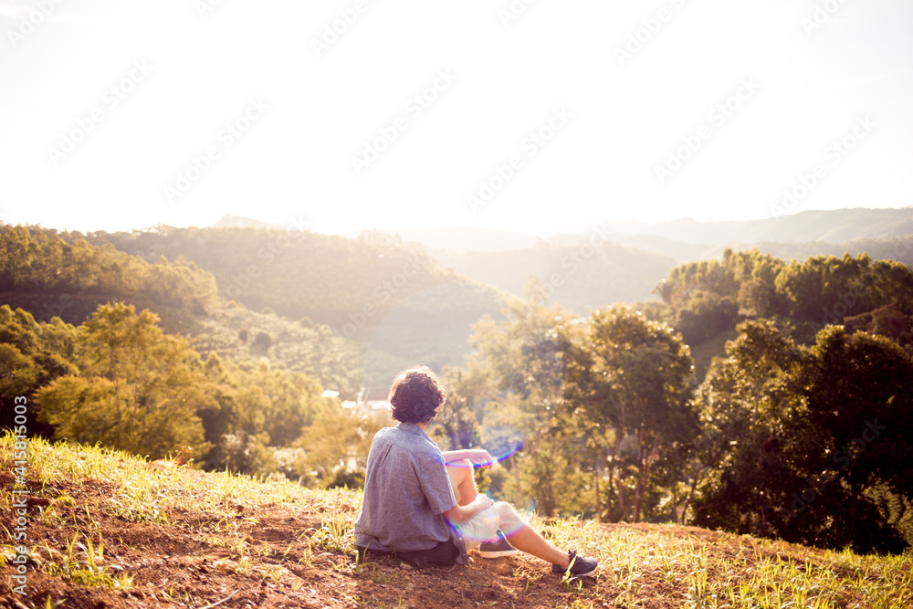 Young man sitting on the top of a hill, watching the sunset in the mountains. Adventure or freedom concept.