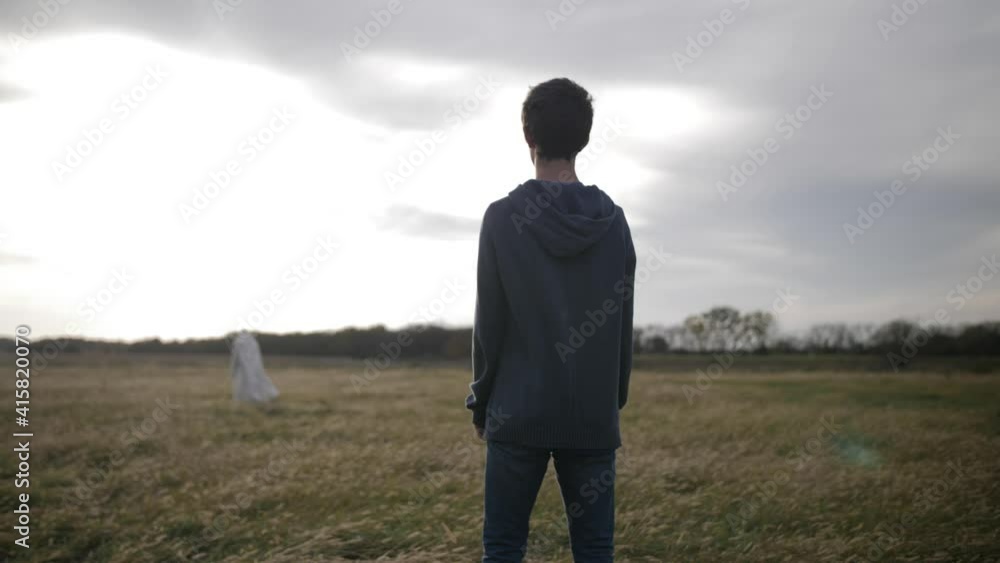 A young teenage boy stands in a field and looks at Jesus Christ - or man wearing a white robe - in the distance with bright sunlight shining. A young teenage boy stands in a field and looks at Jesus Christ - or man wearing a white robe - in the distance with bright sunlight shining.