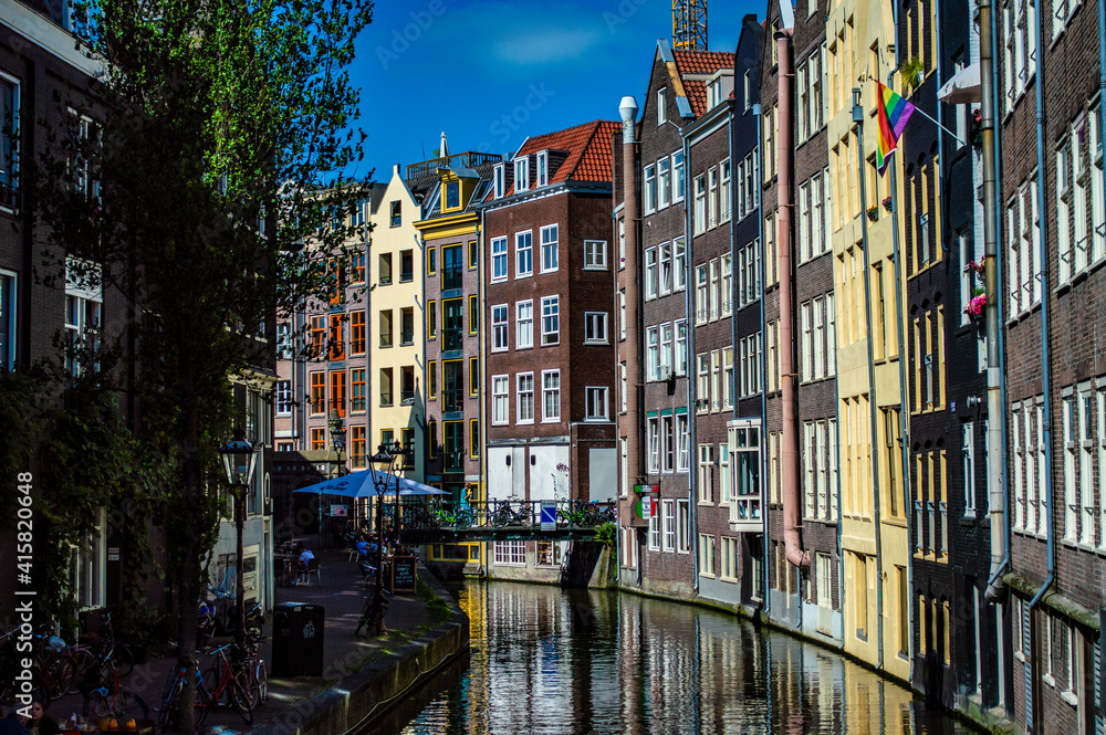 Amsterdam, Netherlands - June 27, 2019: A tranquil canal with a row of traditional brick houses and a rainbow flag in Amsterdam, the Netherlands