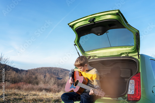Boy 10 years old playing guitar while sitting on the hood of car during a family car trip © olezzo