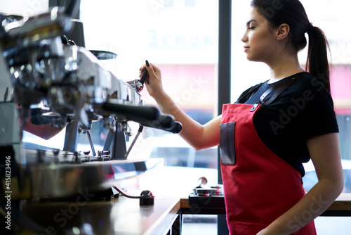 Skilled woman barista using professional inalian equipment for preparing aroma beverages at cafeteria, serious caucasian female waitress in apron checking machine for doing coffee ready for serving photo