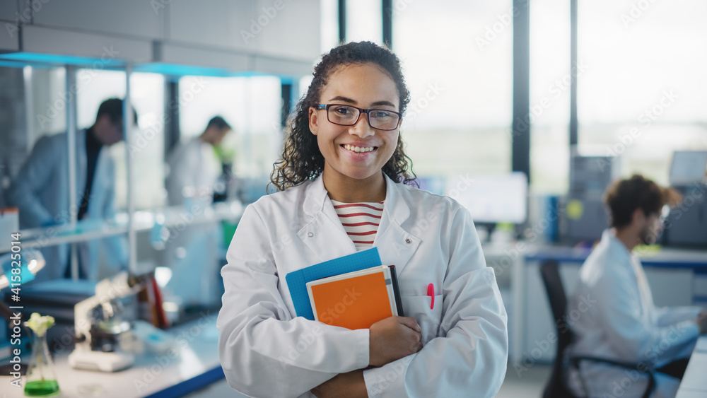 © Gorodenkoff - Medical Science Laboratory: Beautiful Smart Young Black Scientist Wearing White Coat and Glasses, Holds Test Books, Smiles Looking at Camera. Diverse Team of Specialists. Medium Portrait Shot