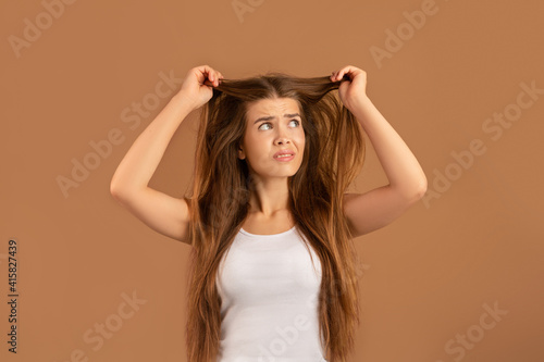 What a disaster. Upset young lady looking at her tangled long straight hair on brown studio background