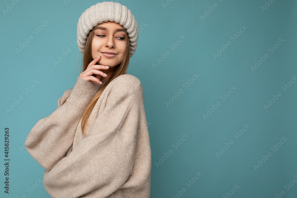 Shot of sexy pretty happy young dark blonde woman standing isolated on blue background wall wearing beige warm sweater and winter beige hat looking down Shot of sexy pretty happy young dark blonde woman standing isolated on blue background wall wearing beige warm sweater and winter beige hat looking down