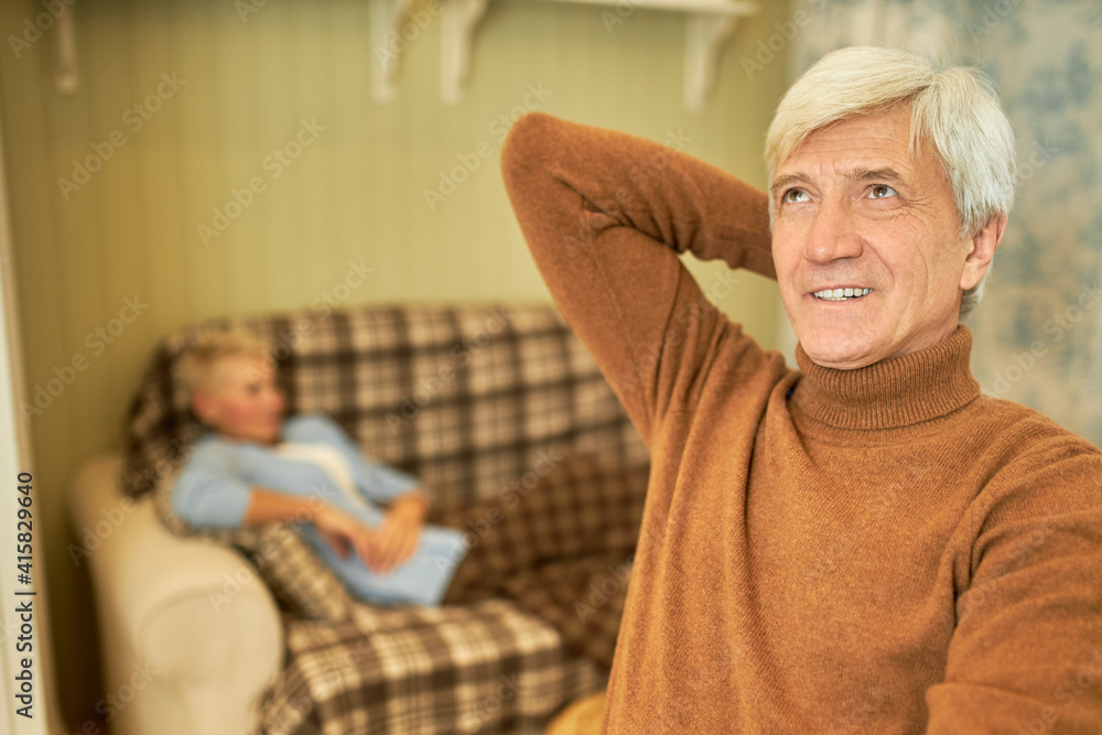 Obraz premium Handsome senior retired man wearing brown turtleneck sweater relaxing at home looking up with thoughtful dreamy smile, his wife having rest on couch in background. Quarantine and social distancing
