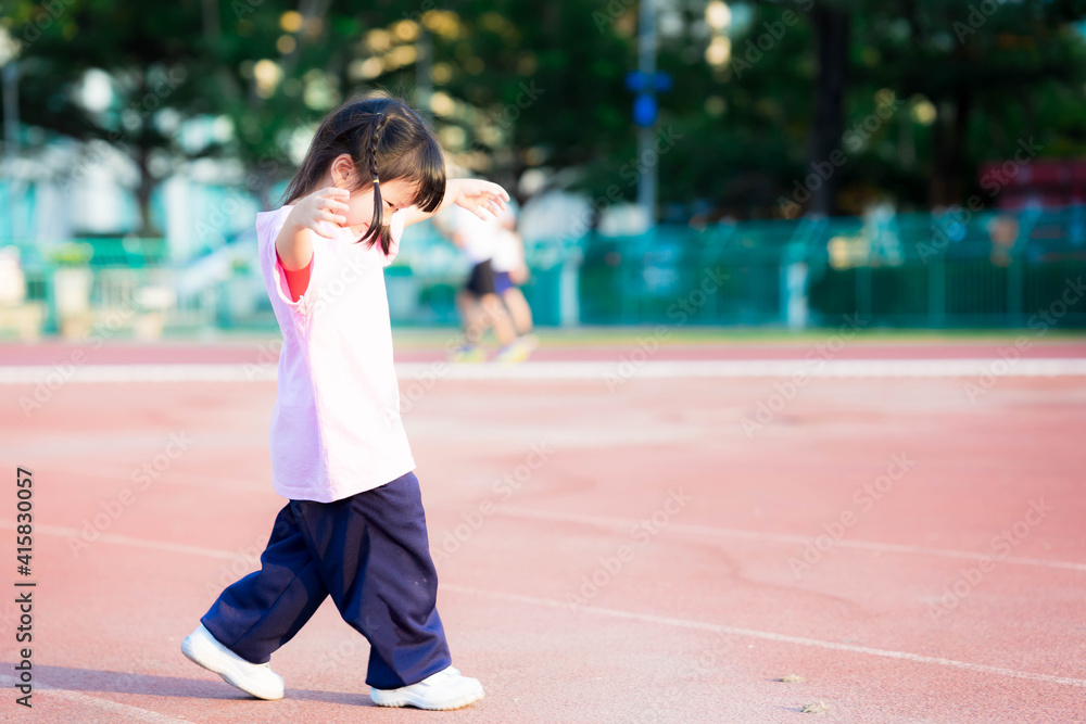 Cute girl with sweet smile looked down at stadium floor. Asian children are exercising. Child enjoy outdoor exercise. Kid play sports with family in public areas. Baby wearing pink shirt is 3-4 years. Cute girl with sweet smile looked down at stadium floor. Asian children are exercising. Child enjoy outdoor exercise. Kid play sports with family in public areas. Baby wearing pink shirt is 3-4 years.