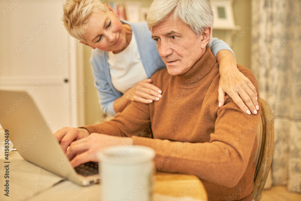 Fototapeta premium Portrait of serious middle aged businessman sitting at table with another cup of coffee, keyboarding on laptop, looking at screen with focused expression, his wife worrying about his health state