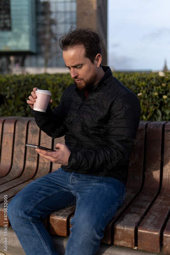 young man sitting on bench in university having coffee and checking his mobile phone young man sitting on bench in university having coffee and checking his mobile phone