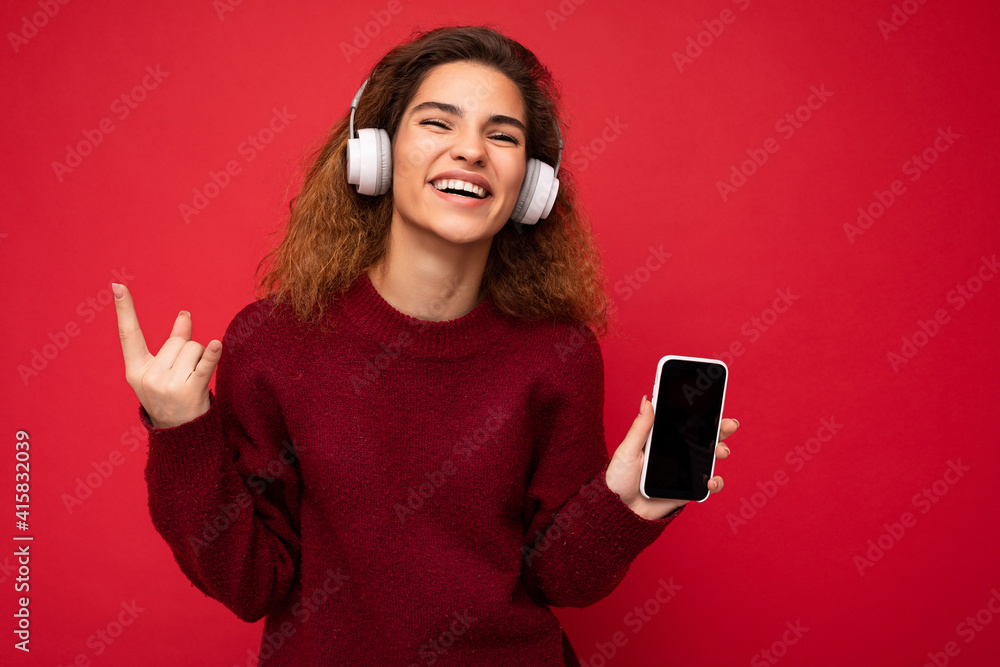 Shot of attractive positive smiling young brunet curly woman wearing dark red sweater isolated on red background wall wearing white wireless headphones and listening to music and showing mobile phone Shot of attractive positive smiling young brunet curly woman wearing dark red sweater isolated on red background wall wearing white wireless headphones and listening to music and showing mobile phone