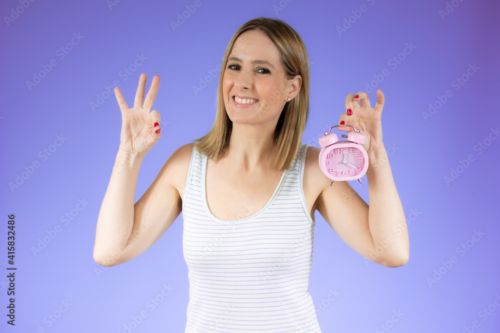Young beautiful smiling woman holding a clock over purple background. Young beautiful smiling woman holding a clock over purple background.