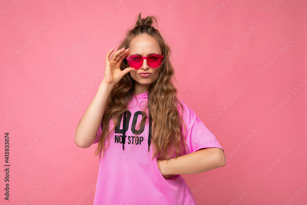 Photo shot of pretty sad dissatisfied young blonde curly woman isolated over pink background wall wearing casual pink t-shirt and stylish colourful sunglasses looking at camera and having questions Photo shot of pretty sad dissatisfied young blonde curly woman isolated over pink background wall wearing casual pink t-shirt and stylish colourful sunglasses looking at camera and having questions