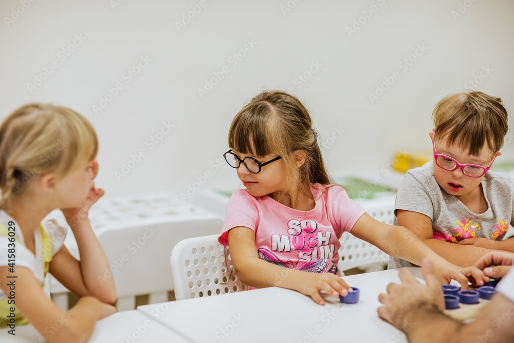 Young kids with down syndrome studying and playing in the room with white desks. Focus is at the girl in the middle. Young kids with down syndrome studying and playing in the room with white desks. Focus is at the girl in the middle.