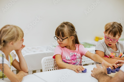 Young kids with down syndrome studying and playing in the room with white desks. Focus is at the girl in the middle.