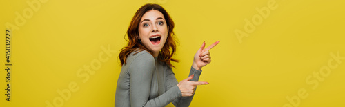 excited woman looking at camera while pointing with fingers isolated on yellow, banner
