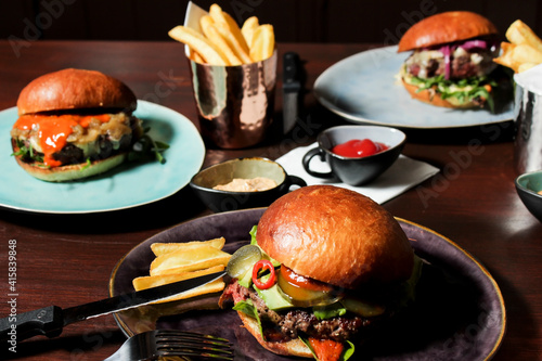 Different burgers on the table, served frenc fries and sauces on plates. The closest spicy beef burger with jalapenos, pickled cucumbers and avocado with spicy sauce on a plate with fork and knife. photo