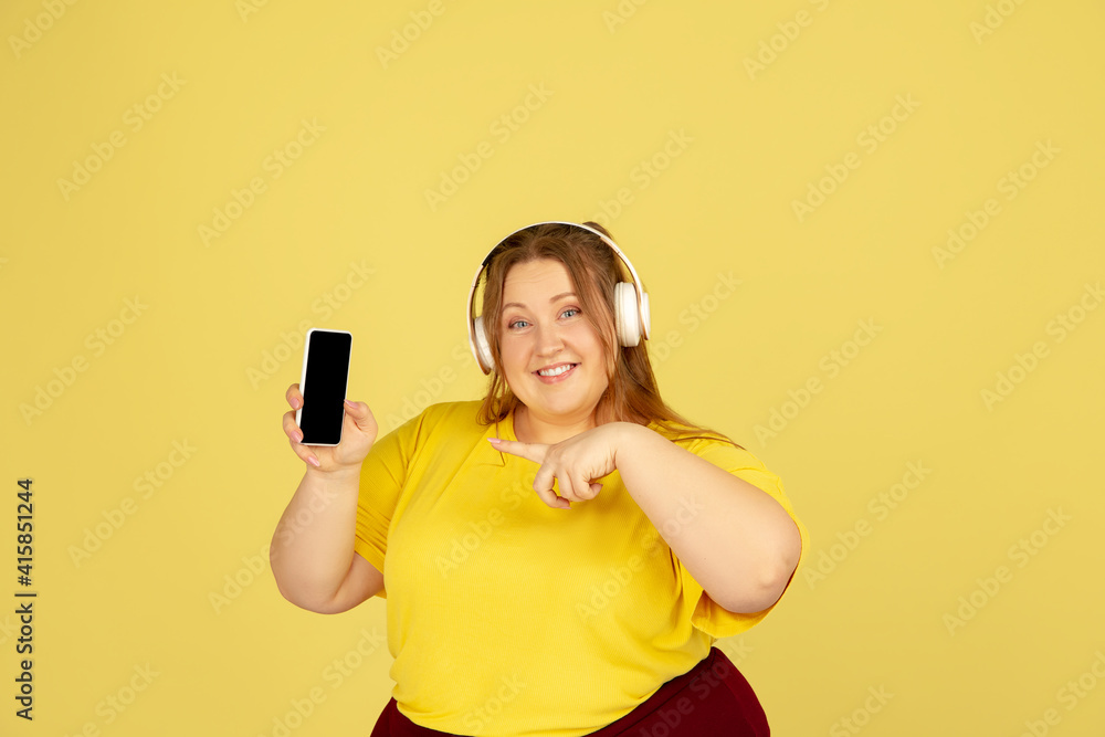 Shows phone screen. Beautiful plump caucasian plus size model isolated on yellow studio background. Concept of inclusion, human emotions, facial expression, sales, body positive. Copyspace for ad. Shows phone screen. Beautiful plump caucasian plus size model isolated on yellow studio background. Concept of inclusion, human emotions, facial expression, sales, body positive. Copyspace for ad.