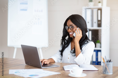 Friendly young black woman in office clothes talking on mobile phone at workplace, empty space