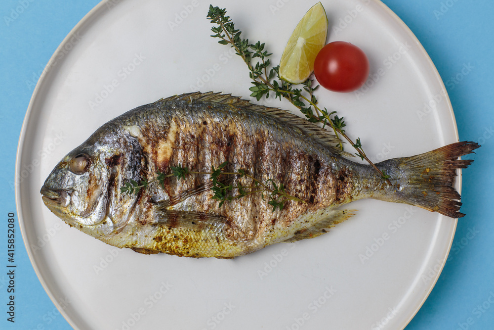 Grilled dorado with thyme, tomato, lemon and spices in a white plate on a blue background Grilled dorado with thyme, tomato, lemon and spices in a white plate on a blue background