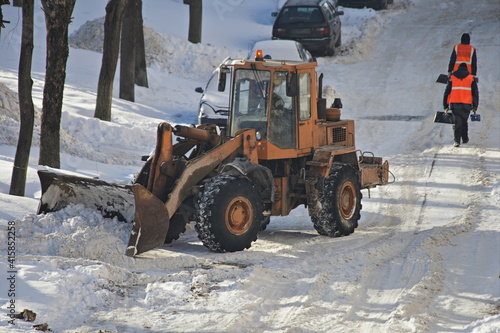 One yellow powerful wheeled tractor removes a snow with scraper shovel blade snowplow on city courtyard road after heavy snowfall at winter day on janitors workers background, top front side view