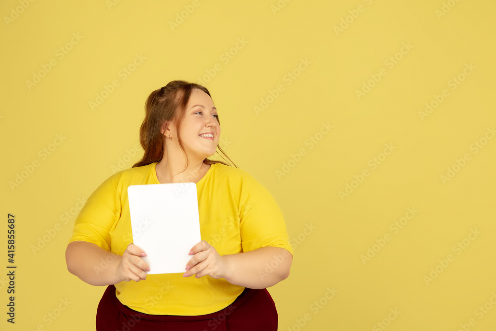Using tablet. Beautiful plump caucasian plus size model isolated on yellow studio background. Concept of inclusion, human emotions, facial expression, sales, body positive. Copyspace for ad. Using tablet. Beautiful plump caucasian plus size model isolated on yellow studio background. Concept of inclusion, human emotions, facial expression, sales, body positive. Copyspace for ad.