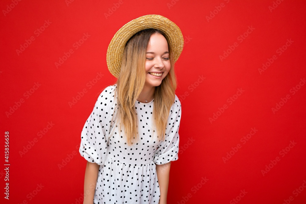 Photo shot of young beautiful cute happy blonde woman wearing casual dress and straw hat isolated over red background with copy space Photo shot of young beautiful cute happy blonde woman wearing casual dress and straw hat isolated over red background with copy space
