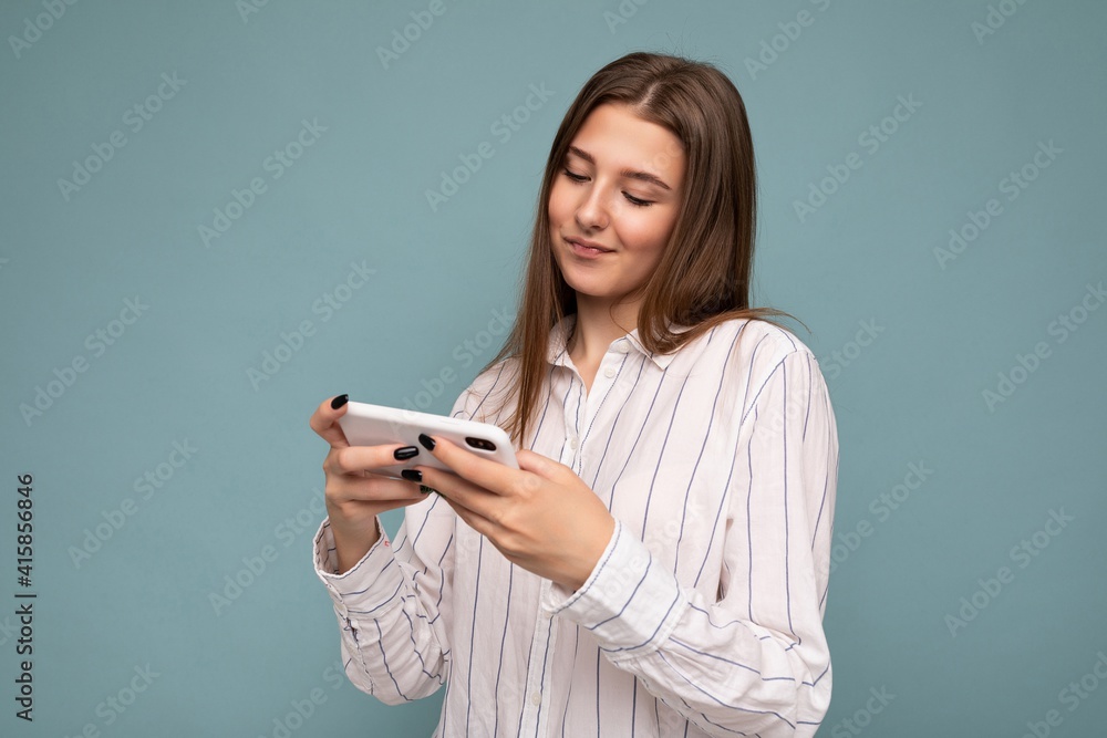 Concentrated happy attractive young blonde woman wearing casual white shirt isolated over blue background wall holding smartphone and playing online games looking at gadjet screen Concentrated happy attractive young blonde woman wearing casual white shirt isolated over blue background wall holding smartphone and playing online games looking at gadjet screen
