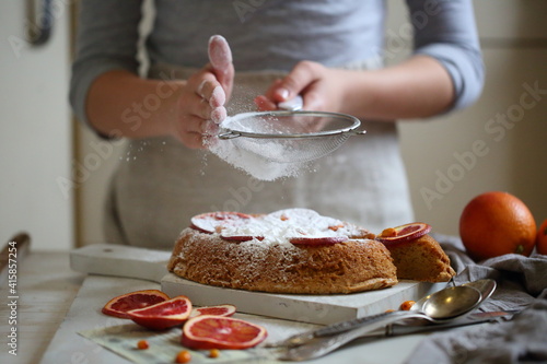Children's hands sprinkle icing sugar through a sieve on a pie with oranges