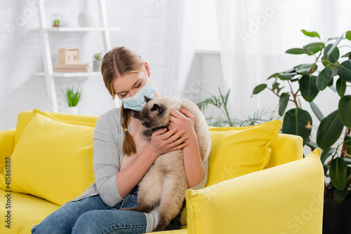 Young woman with allergy reaction wearing medical mask and holding furry cat on yellow couch