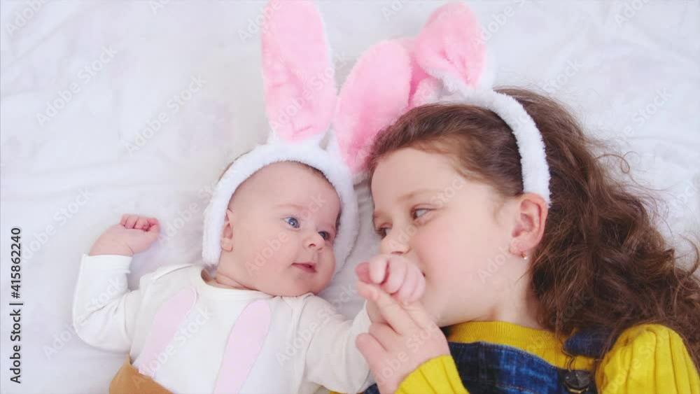 Close up of cute beautiful preschool girl holding little baby sister's hand lying on comfortable white bed, dressed in pink bunny fluffy ears, happily spending time together during Easter celebration Close up of cute beautiful preschool girl holding little baby sister's hand lying on comfortable white bed, dressed in pink bunny fluffy ears, happily spending time together during Easter celebration