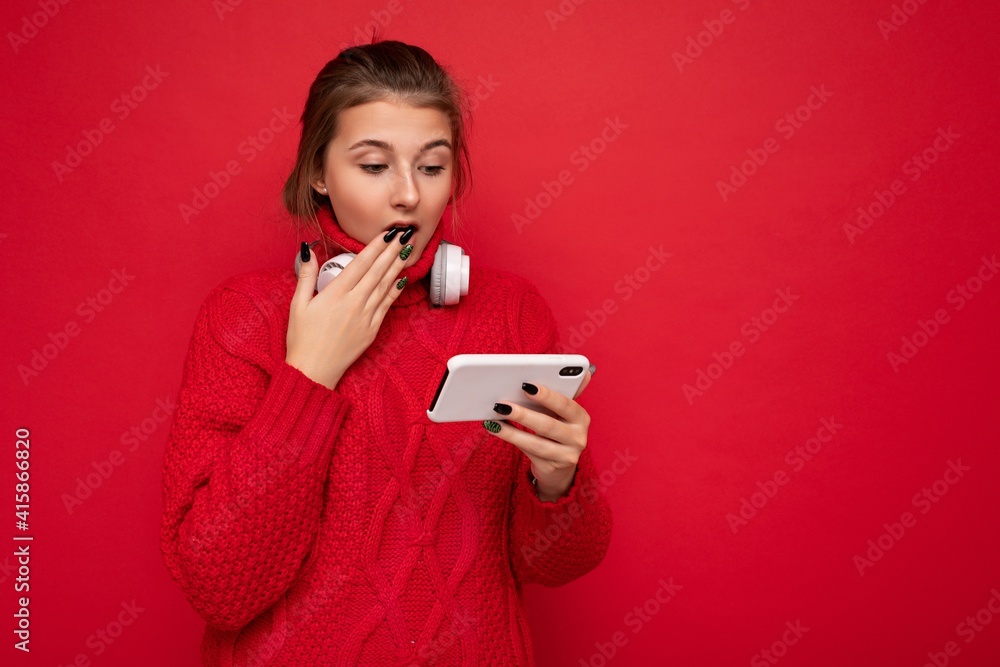 Attractive shocked young brunet woman wearing red sweater isolated over red background wall holding and using mobile phone surfing on the internet and wearing bluetooth headphones looking at Attractive shocked young brunet woman wearing red sweater isolated over red background wall holding and using mobile phone surfing on the internet and wearing bluetooth headphones looking at