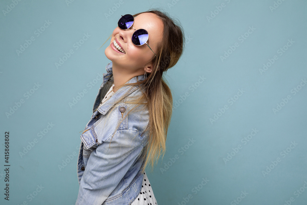Shot of pretty positive smiling young blonde woman wearing blue jean jacket and stylish sunglasses isolated over blue background wall looking to the side Shot of pretty positive smiling young blonde woman wearing blue jean jacket and stylish sunglasses isolated over blue background wall looking to the side