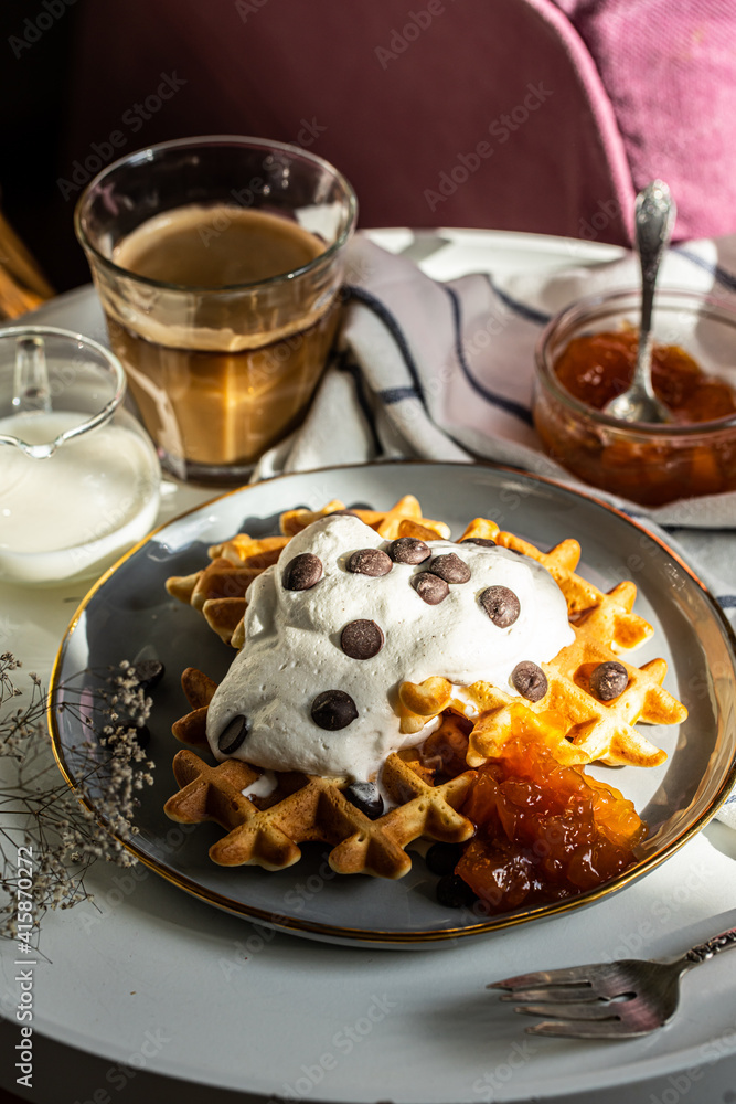 Homemade Belgian waffles with natural whipped cream, chocolate droplets and apricot jam, glass of coffee , milk jug and jam on little round table near armchair . Homemade Belgian waffles with natural whipped cream, chocolate droplets and apricot jam, glass of coffee , milk jug and jam on little round table near armchair .