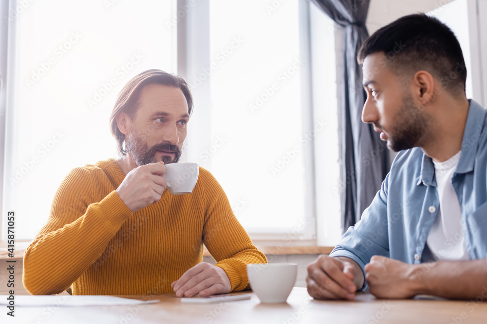 Bearded man holding coffee cup during conversation with Hispanic son in kitchen