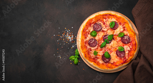 Delicious pepperoni pizza, spices, basil and linen napkin on a brown background. Top view, panorama.