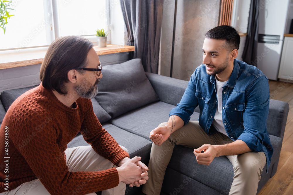 Fototapeta premium Young Hispanic man gesturing while talking to dad on sofa at home