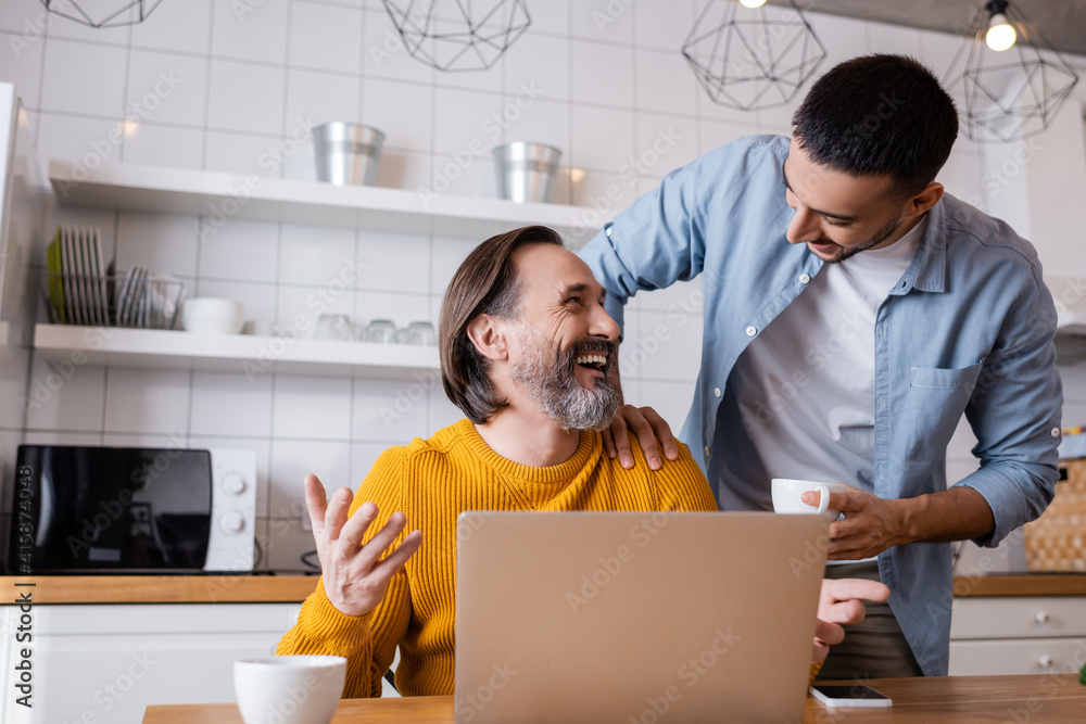 Laughing man looking at young Hispanic son while sitting near laptop in kitchen Laughing man looking at young Hispanic son while sitting near laptop in kitchen
