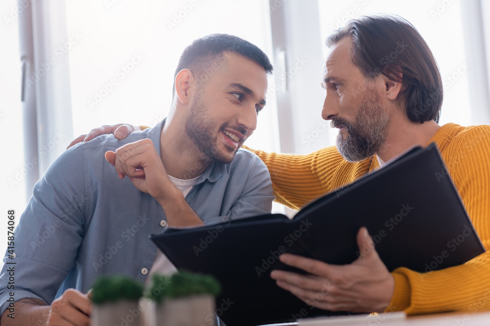 Smiling Hispanic man looking at father holding photo album on blurred foreground Smiling Hispanic man looking at father holding photo album on blurred foreground