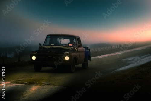 antigua pickup deteriorada circula por un camino de tierra  en un clima nublado y brumoso  con dos personas en su interior  mientras cae el sol