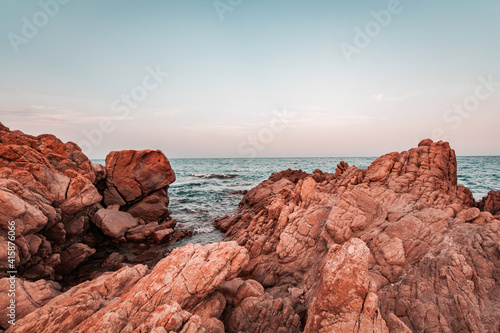 Beautiful sunset seascape in Italy, Sardinia island. Red and pink rocks, wind erosion, geology stratification, air oxidation. Teal and orange lighting, endless horizon and soft pastel colored clouds.