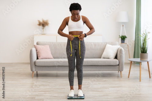 Tableau sur toile Shocked Black Woman Weighing Herself And Measuring Waist At Home