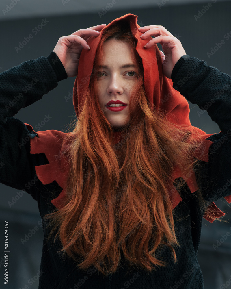 Portrait of beautiful red-haired young woman in hood looking at camera. Girl in the image of Red woman.