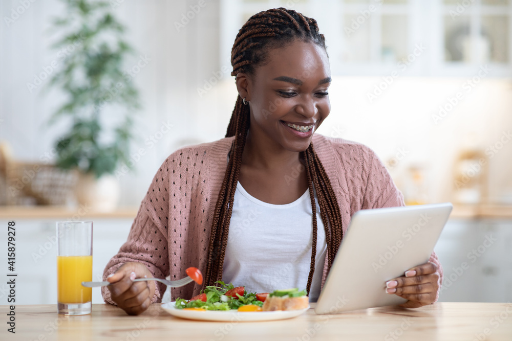 Cheerful Black Woman With Digital Tablet Reading News During Breakfast In Kitchen