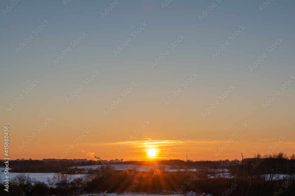 Winter evening landscape with sunset and multicolor sky. A view of the snow-covered area with dry grass, trees and bushes.