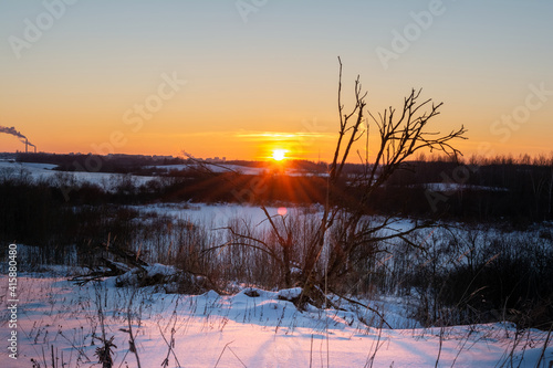 A view of the snow-covered area with dry grass, trees and bushes. On the horizon is an urban industrial area with smoking chimneys. Winter evening landscape with sunset