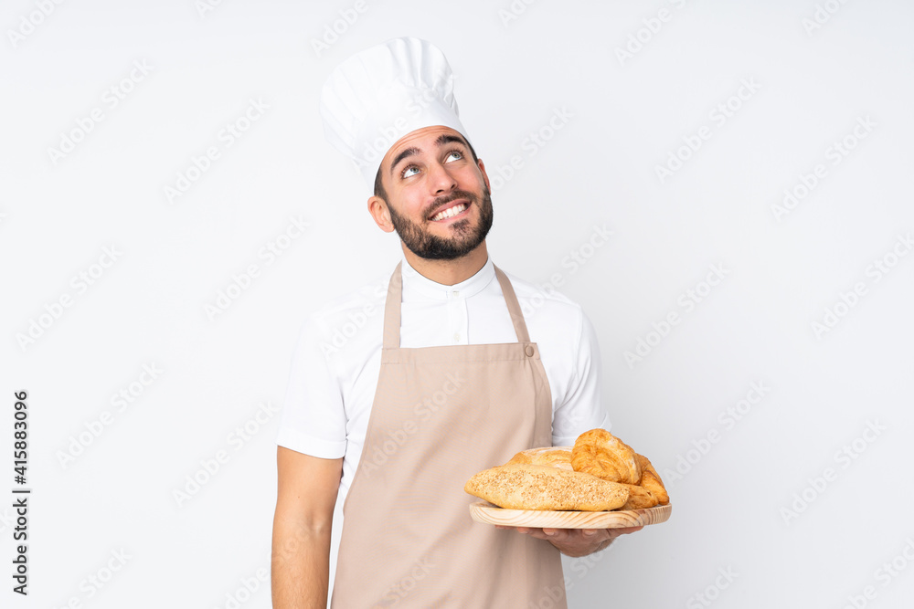 Male baker holding a table with several breads isolated on white background laughing and looking up