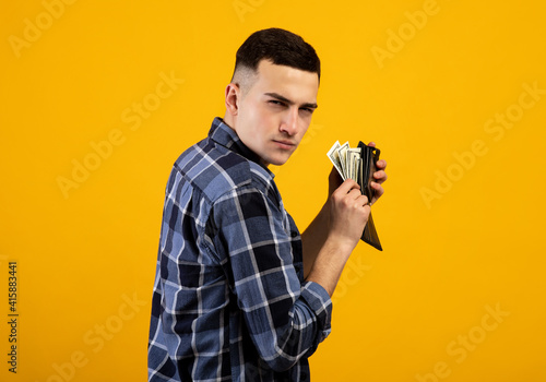 Young man with wallet full of money being greedy over his wealth on orange studio background photo