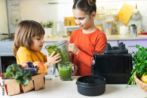 Children drink green smoothies from a blender in the kitchen. Funny Brother and Sister Cook Healthy Breakfast together. Cute kids learn Healthy Habits and how to make green cocktail